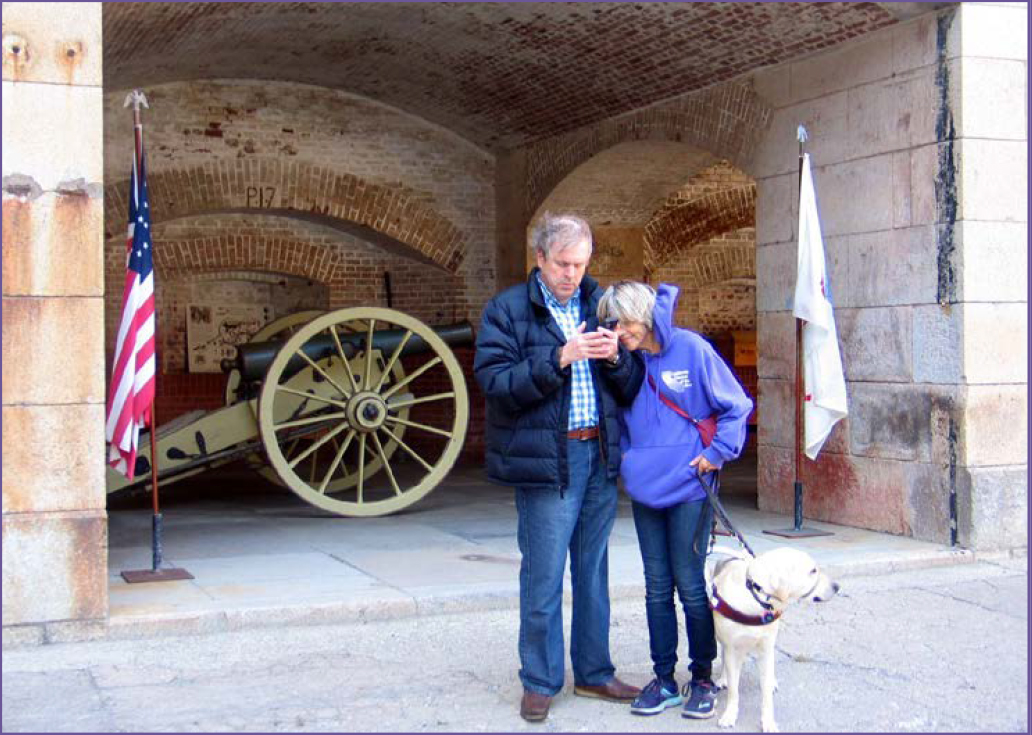 California Council of the Blind members Susan and John Glass stand in front of a brick casement housing a civil war cannon as theyem listen to audio description of Fort Point on the UniD app.