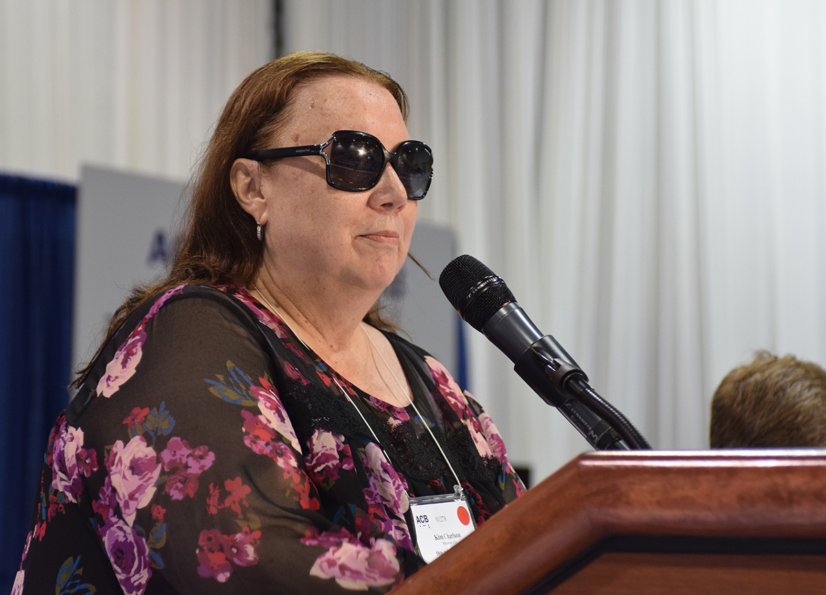 ACB president Kim Charlson reviews the highlights of her six years in office. She stands behind the lectern microphone, sporting a black dress with a pink floral print.