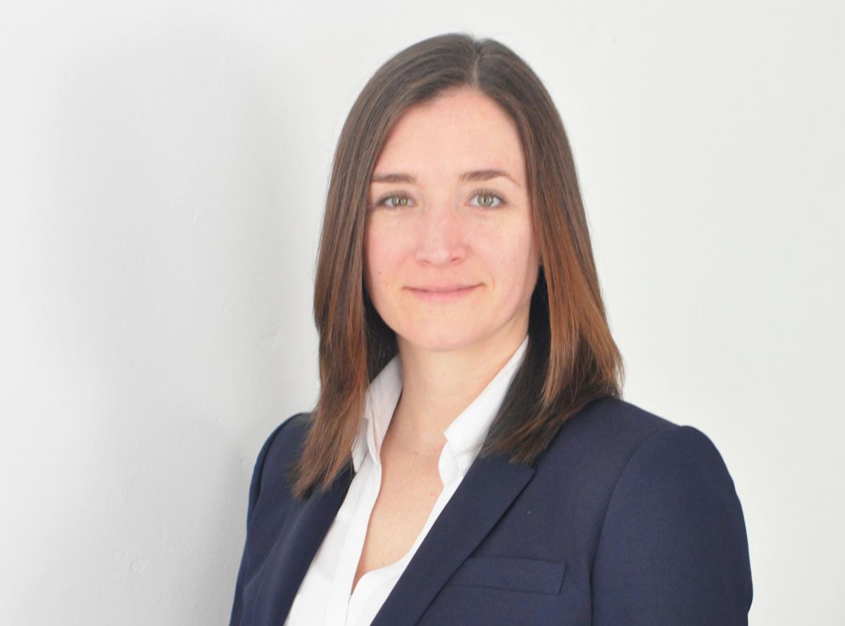 Claire Blome, a white woman with straight, fine brown hair just past her shoulders wears a navy suit jacket and white blouse.