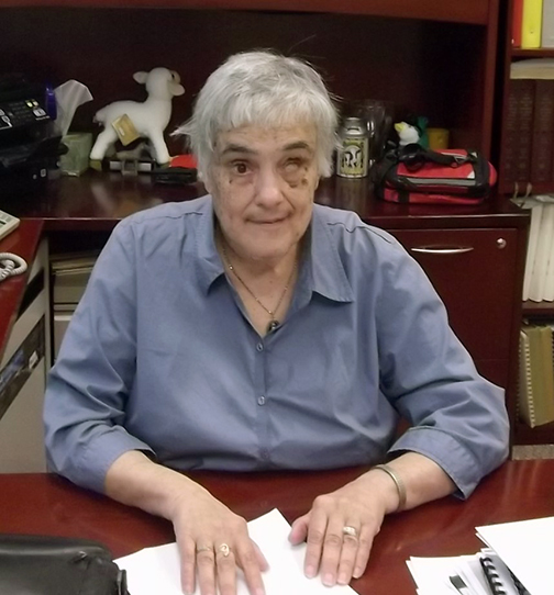 Camille sits at a desk, wearing a blue button up shirt, reading a braille document.