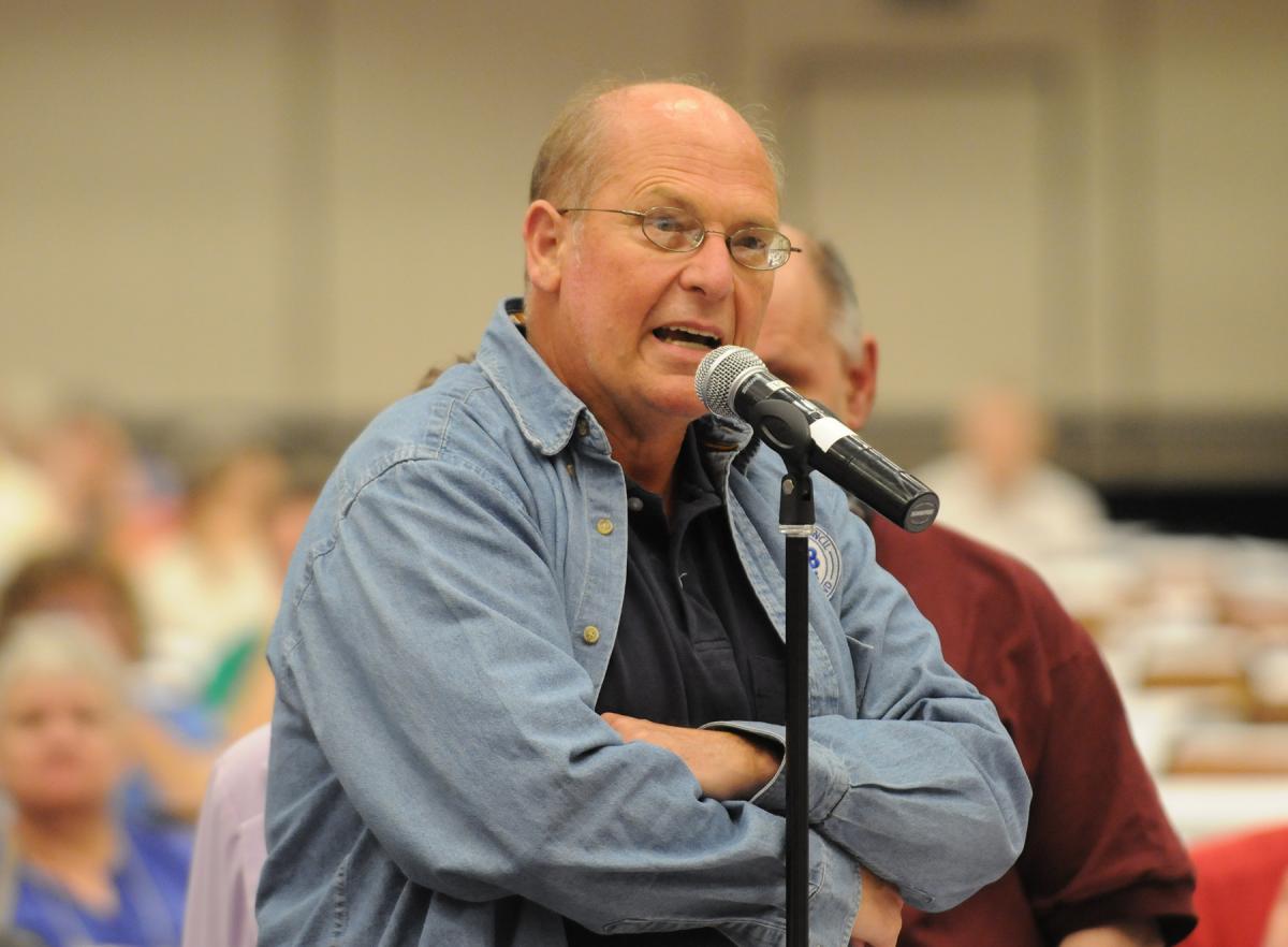 Wearing a blue button up shirt, Rod speaks into a microphone in front of an audience at convention, arms crossed. 