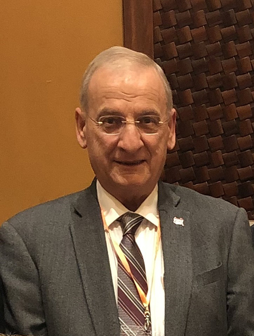 Dan, wearing a grey suit, striped tie, and glasses, smiles as he faces towards the camera.