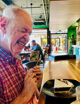 Don sits in front of a piece of pie, grasping a fork with both hands, his face lit up with a big smile.