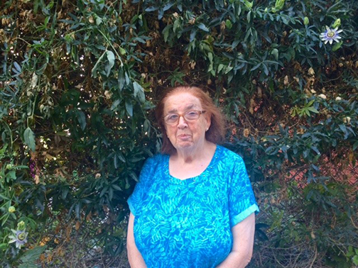 Liz, wearing glasses and a bright blue patterned shirt, stands outside with a wall of purple passion flower vines behind her.