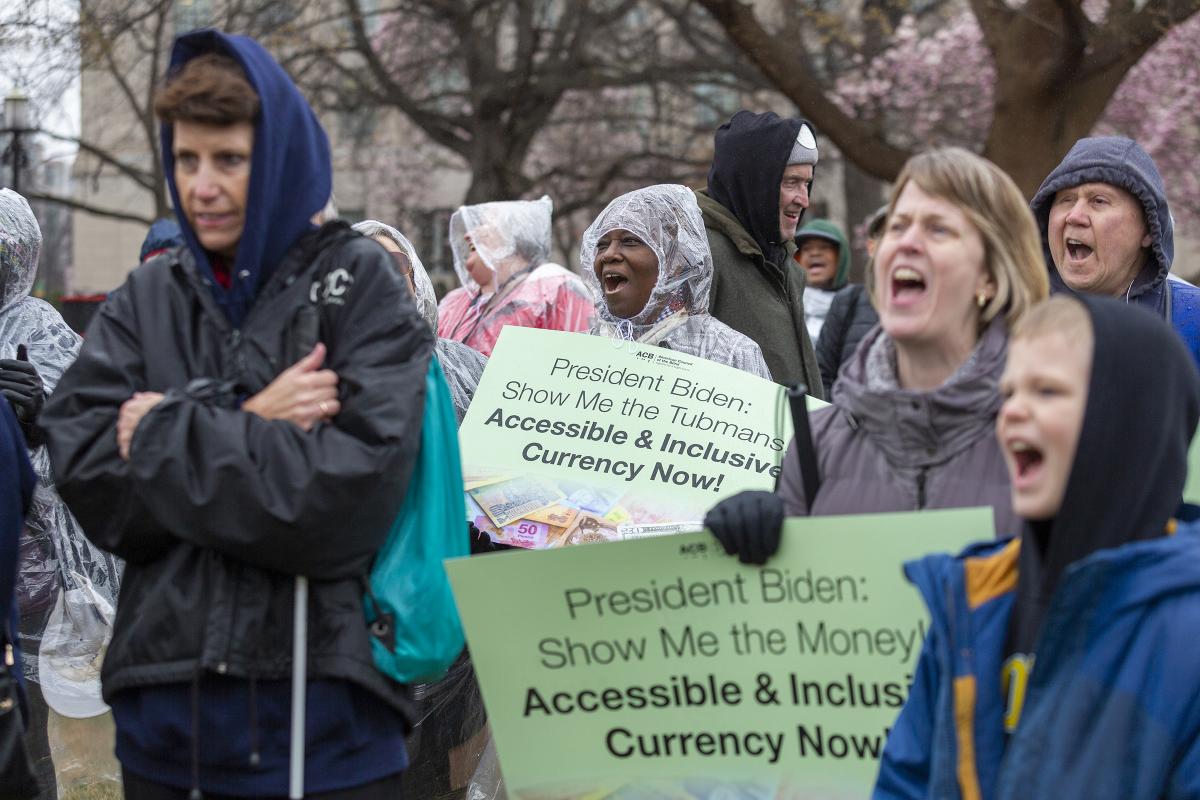 Blind, low vision, and sighted rally participants of all ages shout passionately in the rain as they hold signs in support of accessible and inclusive currency. 