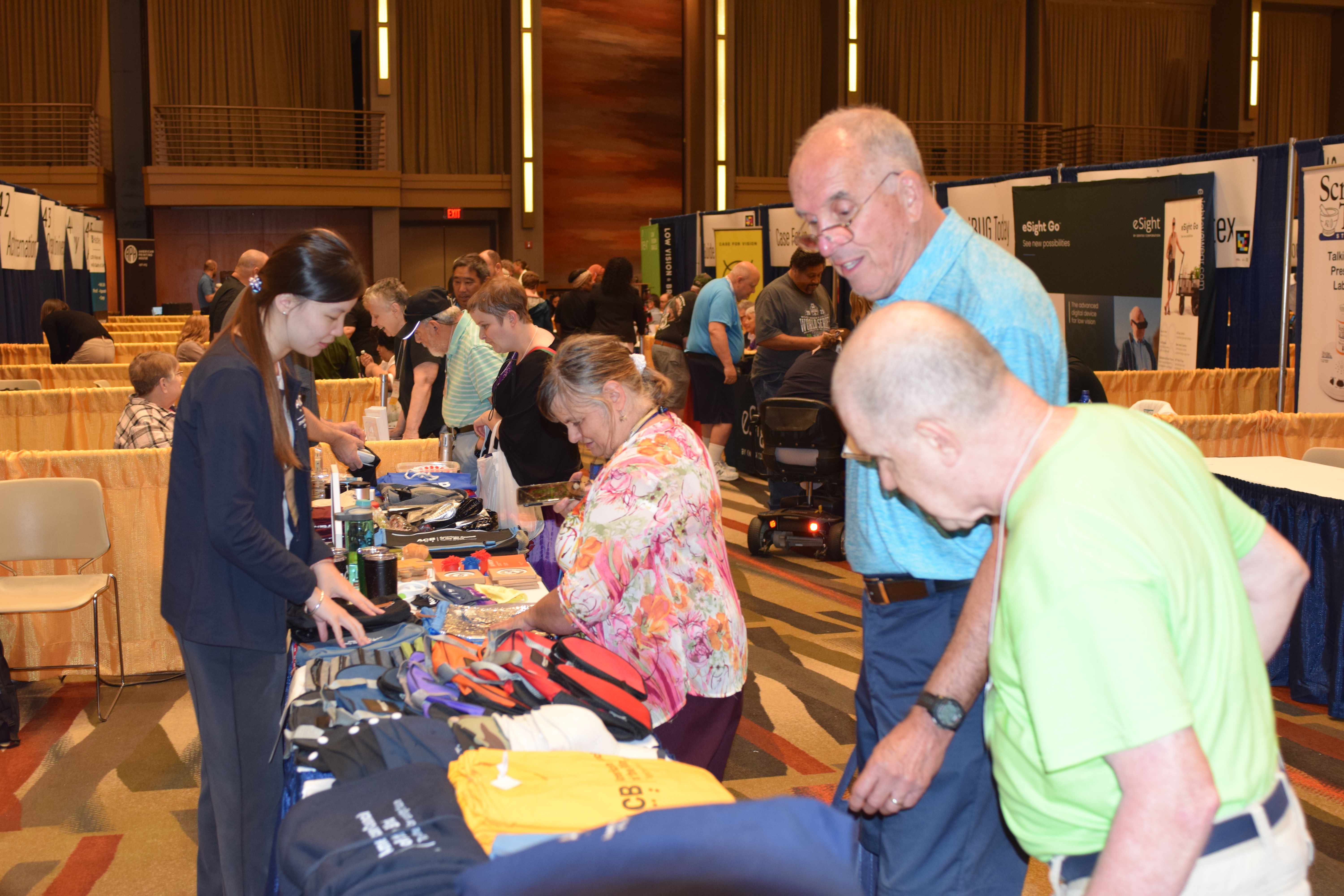 An ACB staff member in a dark blue cardigan behind a table for the mini mall booth helping several members out in the exhibit hall. The table has an assortment of items including hoodies, t-shirts, water bottles, canes, and bags. Behind them are dozens of other booths with dozens of other members and people.
