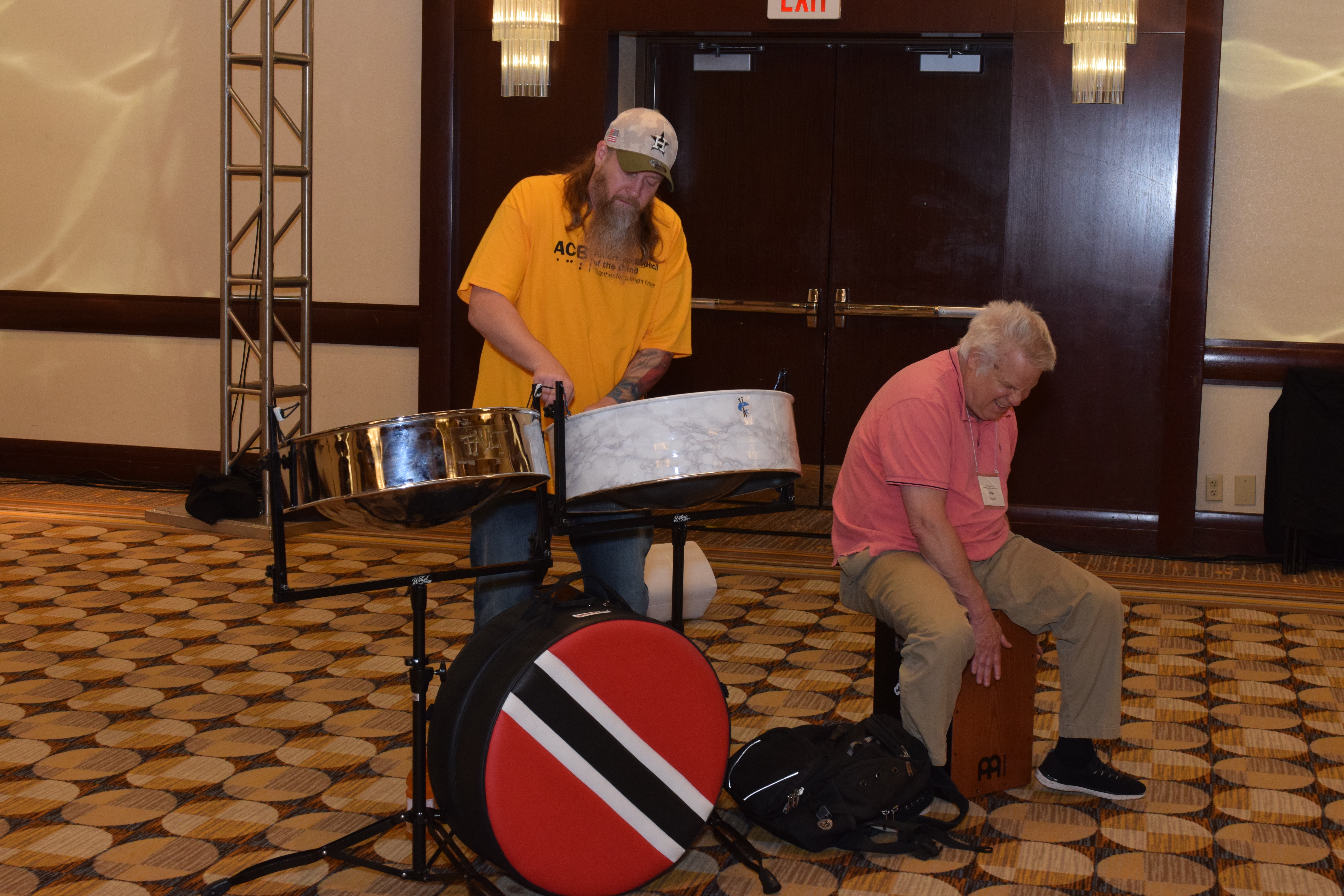 2025 scholarship winner Frank Ritter wearing a mustard yellow ACB t-shirt on the steel drums. Frank's drums are steel, white marbled, and red with a white and black stripe. Seated on his lower right is Peter Altschul on wooden drums.