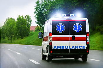 A photo of an ambulance car with the sirens flashing white as it travels on a main road with trees and grass on the right side of the car. The ambulance car is white with a singular red stripe all around. On the backdoors of the car there are two blue medical symbols with the words "Ambulance" printed on the bottom. (Photo via Canva)