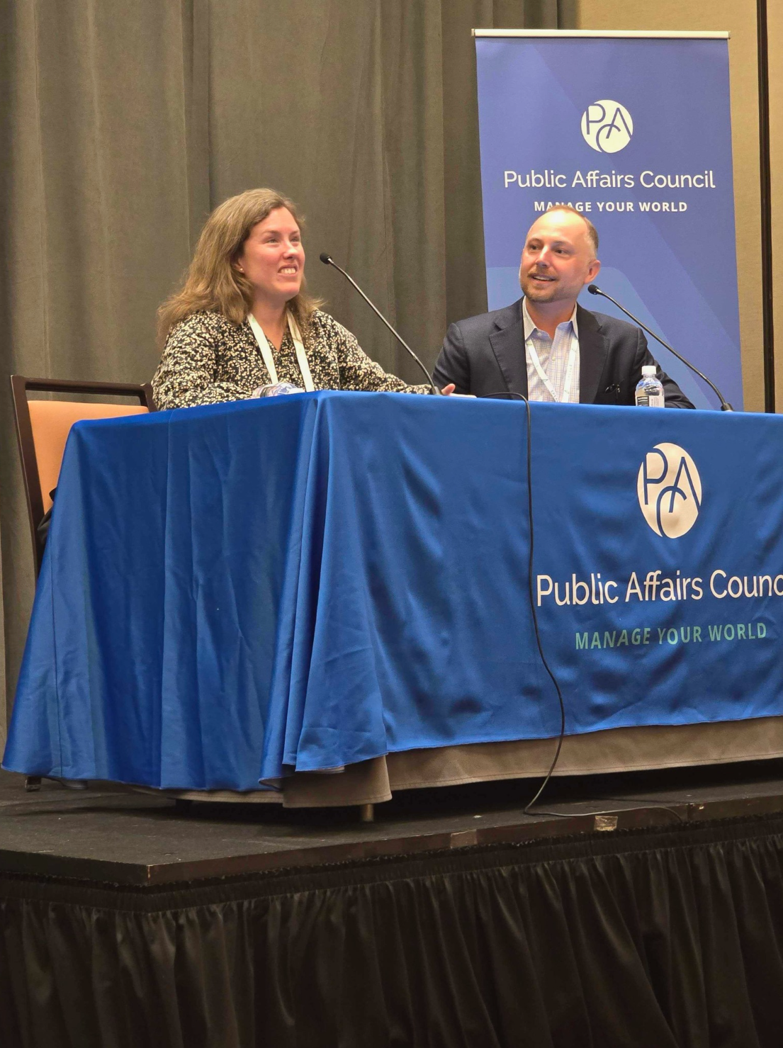 Kolby Garrison, ACCS Coordinator (left) and Tyler Hargrave from Speak4 (right) speaking on stage at a table. Behind them is a banner displaying the Public Affairs Council logo and the text, “Public Affairs Council, Manage Your World