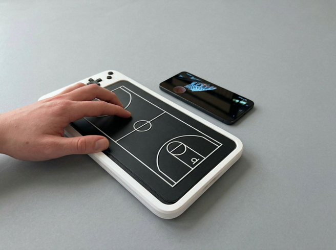 A stock image of a hand using the OneCourt's haptic tablet. The haptic tablet is rectangular shaped. The outer rim is white with a black screen and black buttons on the far right. The screen is replicating a basketball court. There is a black smartphone placed in front of the haptic tablet that showcases a basketball game.