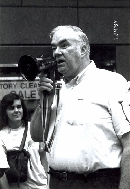 Oral Miller speaks into the megaphone, chanting about “access and safety” on Metrorail, at a rally held July 24, 1994 in front of a Metro station. In the background, leaning against a wall, is Jane Carona.