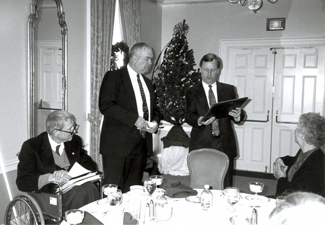 Oral Miller reads the inscription on the plaque that another man is holding before presenting Justin Dart Jr. with the first ACB life membership. Roberta Douglas turns her head toward the men.