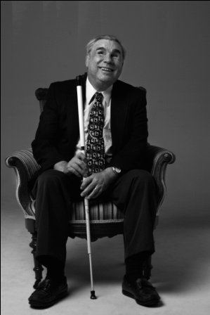 Dr. Ronald E. Milliman smiles for the camera. He sports a dark suit, white shirt, and multicolored tie, and holds onto his cane with both hands.