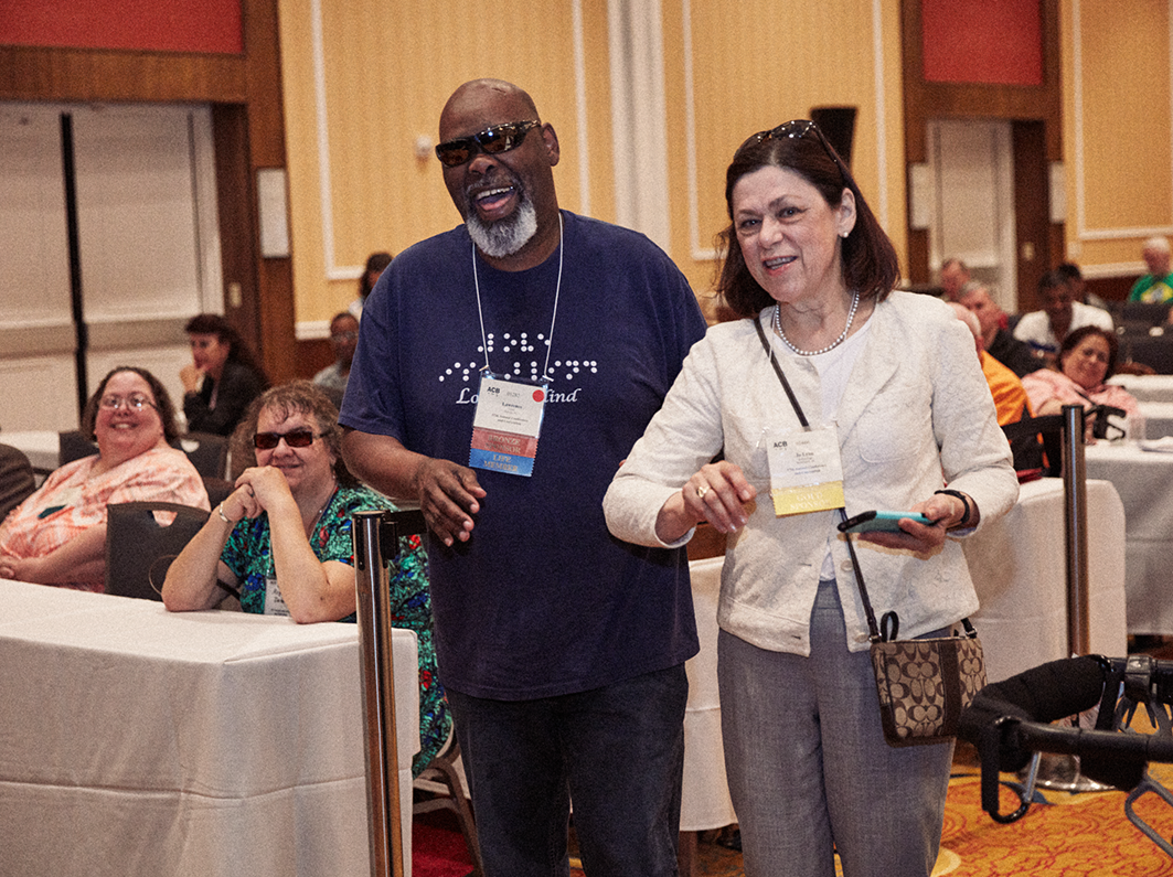 An ACB member smiles and laughs as he walks down the aisle during general session with an ACB staff member.