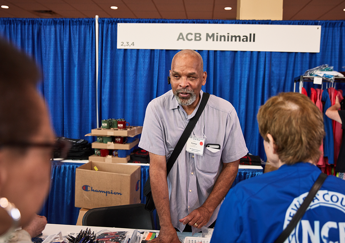An ACB volunteer speaks to ACB members at the ACB Mini Mall booth in the convention Exhibit Hall.